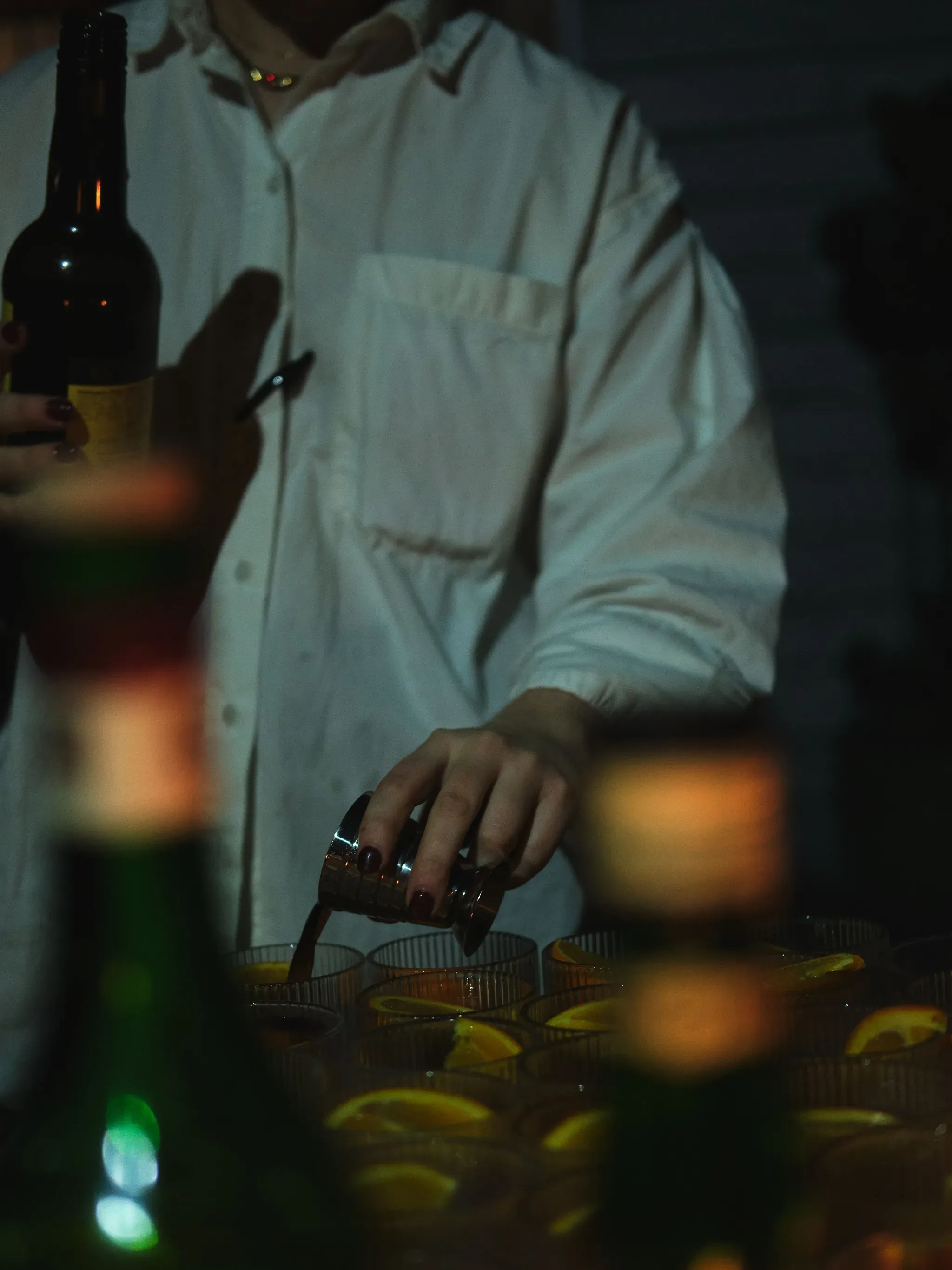 A bartender in a white shirt holding up a dark bottle, green bottles and a tray of glasses blurred in front
